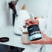 A person in a white sleeve preparing a hot drink with a kettle, a beige mug, and a black jar labeled 'adrenal' on a kitchen counter, indicating the making of a hot beverage.