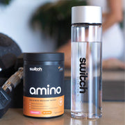 A close-up of a Switch Nutrition amino acid supplement tub with 'EAA & BCAA RECOVERY MATRIX' label next to a branded water bottle on a gym bench, with weights in the background, indicating a focus on fitness and recovery.