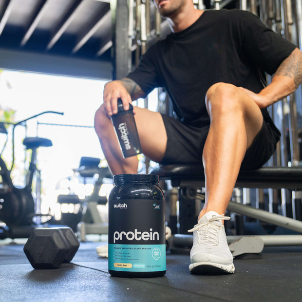 Man in gym gear sitting next to a black Switch Nutrition Protein container on the floor, with gym equipment in the background.