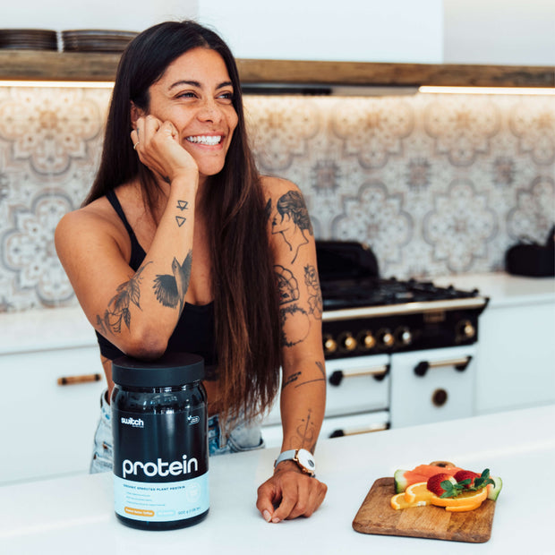 Happy woman with tattoos standing in a kitchen, leaning on a counter with a container of Switch Nutrition Protein, with a plate of fruit in front of her.