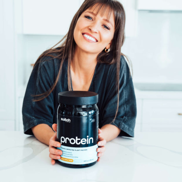Cheerful woman in a casual outfit holding a container of Switch Nutrition Protein in a modern kitchen setting.