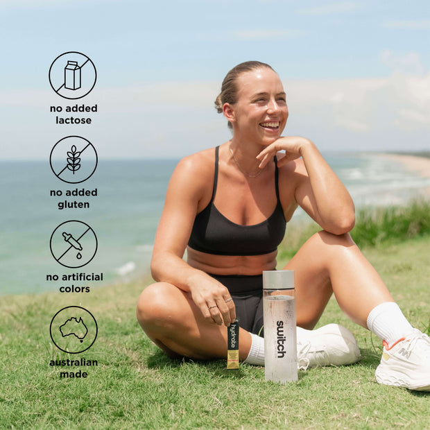 Smiling woman seated on grass by the beach with Switch Nutrition products, emphasising no lactose, gluten, artificial colours, and Australian made.