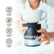 A close-up of a woman's hands holding a striped mug on top of a black jar labeled 'adrenal', with icons indicating 'no added lactose', 'no added gluten', 'no artificial colours', and a badge stating 'Australian made'.