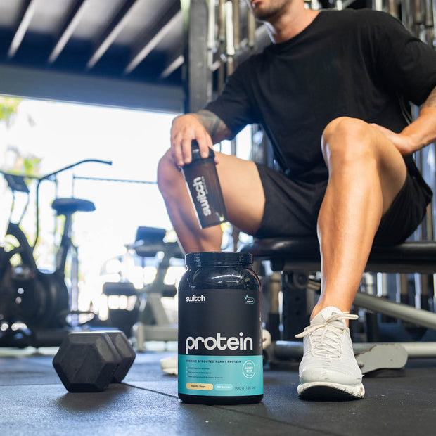 Man in gym gear sitting next to a black Switch Nutrition Protein container on the floor, with gym equipment in the background.