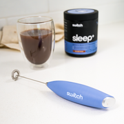 Switch Nutrition Whiz Stick electric mixer in purple, placed on a countertop with a glass of mixed Sleep+ supplement and its container in the background.