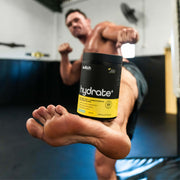 A focused image of 'HYDRATE+' by Switch Nutrition in a gym setting, with a blurred male athlete in the background performing a punch. The product is central and sharp in the foreground, implying its significance in the athlete's training regime. The scene conveys motion and intensity, associating the product with high-energy workouts and athletic performance.