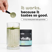 A hand holds a spoonful of green powder over a glass filled with a green smoothie, beside a container of Vitality Switch Greens Powder with the tagline 'it works. because it tastes so good. 30-day money-back taste guarantee.