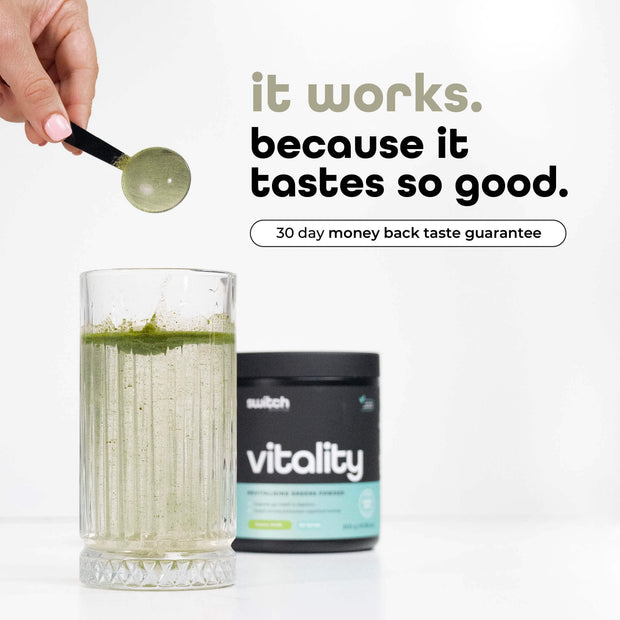 A hand holds a spoonful of green powder over a glass filled with a green smoothie, beside a container of Vitality Switch Greens Powder with the tagline &