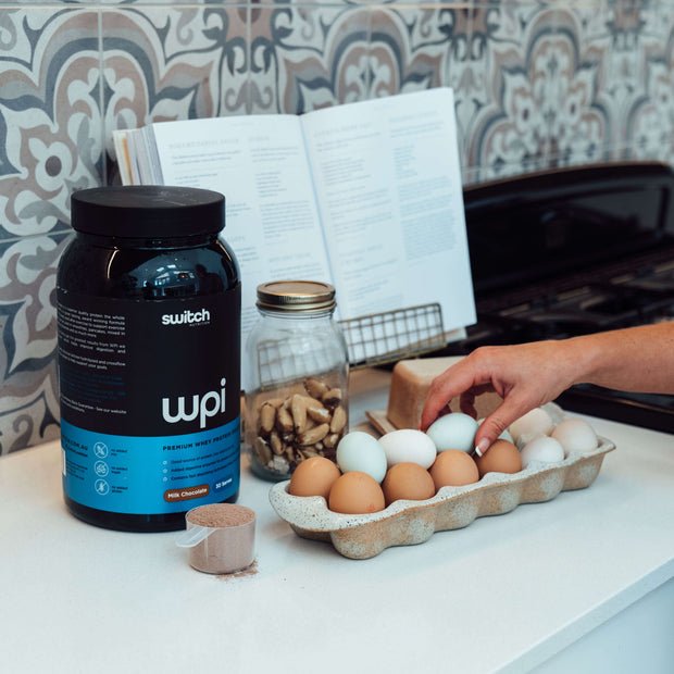 A kitchen scene with a container of WPI Switch Nutrition premium whey protein on the counter, alongside a tray of eggs, a jar of nuts, a cookbook open on a stand, and a person&