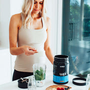 A woman in a light tank top is measuring a scoop of WPI Switch Nutrition whey protein beside a blender filled with spinach and yoghurt, with fresh berries and a jar of milk on the kitchen counter, indicating a health-conscious lifestyle with a focus on nutrition